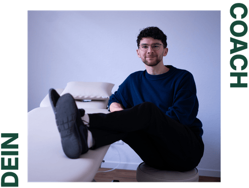 Jonas Ferenc Kohlhage, der Inhaber der Praxis sitzt neben einer Therapiebank und die Beine entspannt auf diese abgelegt. Er lächelt in die Kamera.