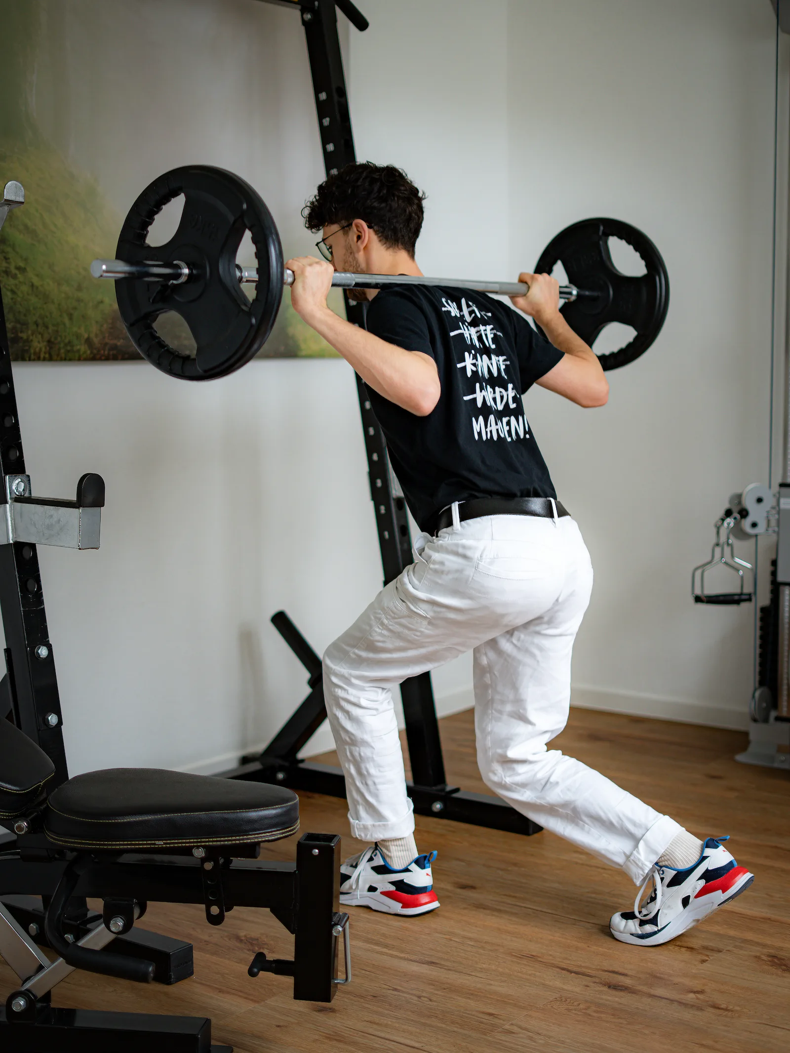 Ein Patient macht im Rahmen der Krankengymnastik am Gerät (KGG) einen Lunge mit einer Langhantel und Gewicht auf dem Rücken. Auf seinem T-Shirt steh "Machen"!