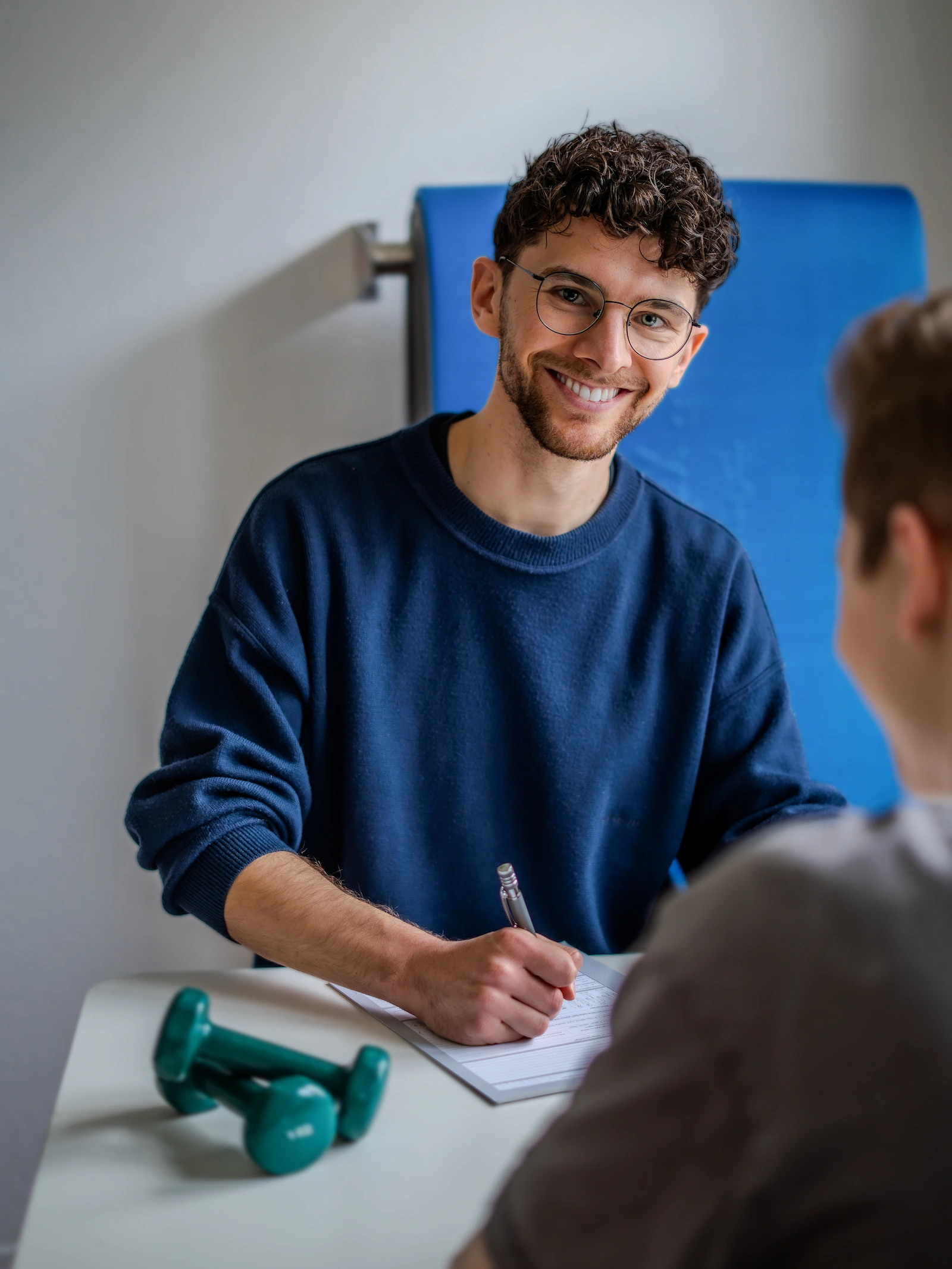 Therapeut sitzt vor einem Patienten und macht sich Notizen zu dem physiotherapeutischen Befund. Im Hintergrund sind Sportmatten zu sehen und auf dem Tisch zwei kleine Hanteln.